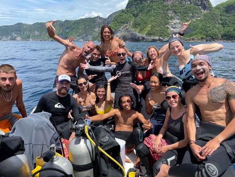 A group of smiling people in bathing suits sit in a boat with green cliffs behind them.